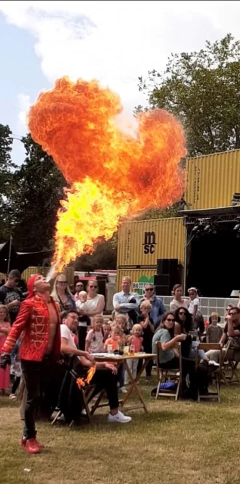 Fire breather Nuno in a red circus jacket blowing a fireball above the crowd at a summer family festival