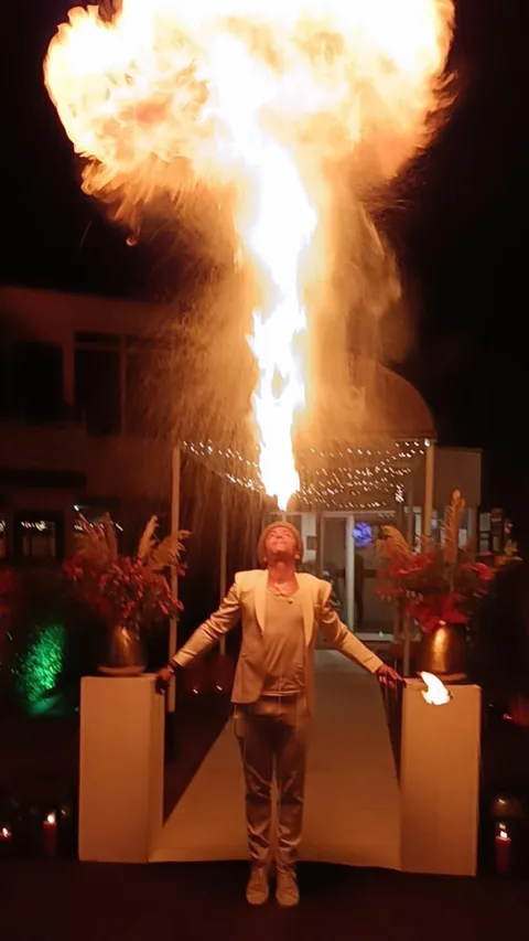 In a white gala suit, Nuno blows a towering column of fire between flowers and candles at an elegant party